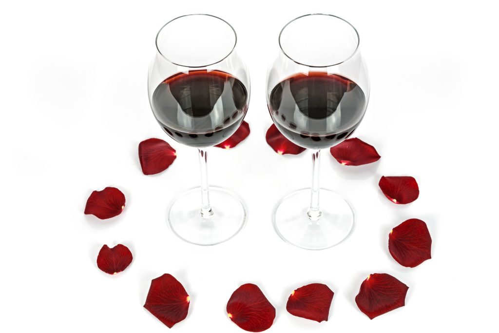 Two wine glasses with red wine surrounded by rose petals on a white background